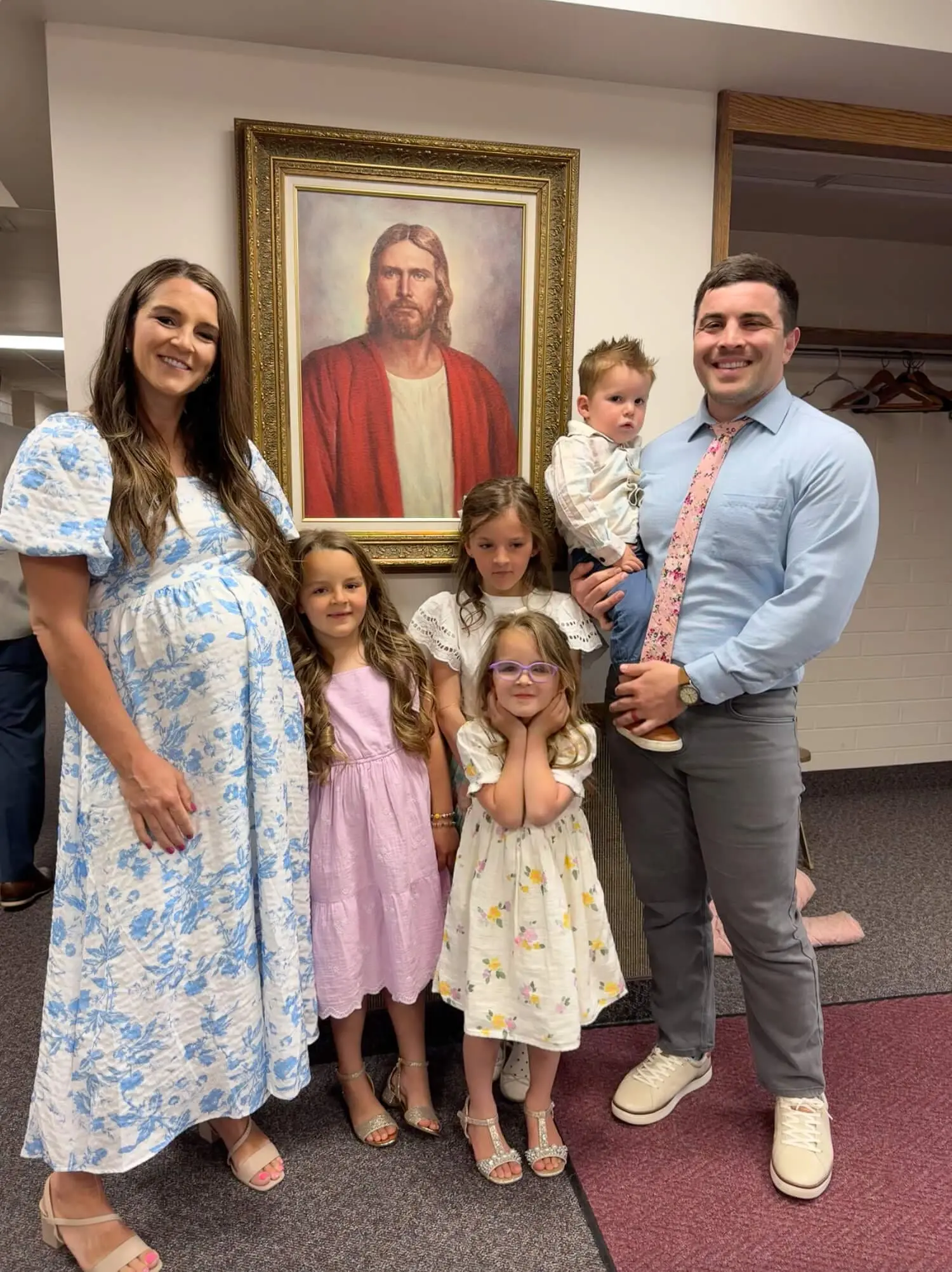 Smiling family of six dressed in Sunday best, standing together in front of a framed portrait of Jesus in a church hallway.